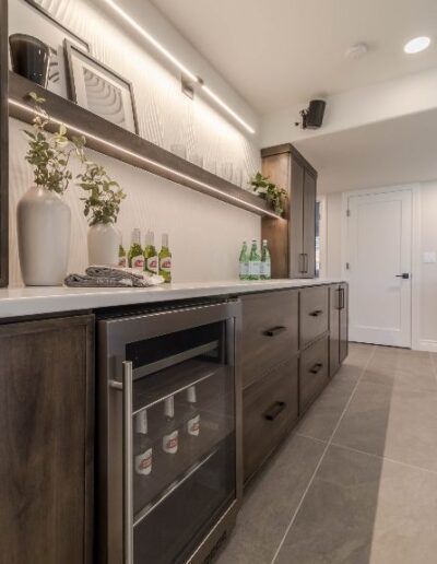 Modern kitchenette featuring a built-in wine cooler, wooden cabinets, under-cabinet lighting, and decorative vases on a white countertop.