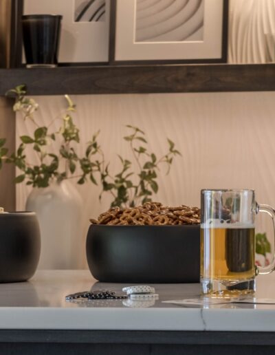 A modern home bar setting with two glasses of beer, bowls of pretzels and popcorn, and decorative plants and artwork in the background.