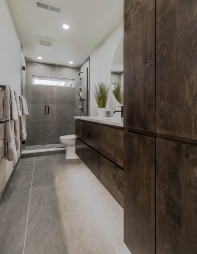 Modern bathroom with dark wood vanity, walk-in shower, and white fixtures, accented with gray tiles and green plants.