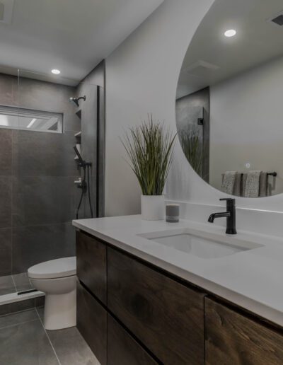 Modern bathroom with large mirror, wooden vanity, white sink, walk-in shower with glass door, and a toilet to the left.