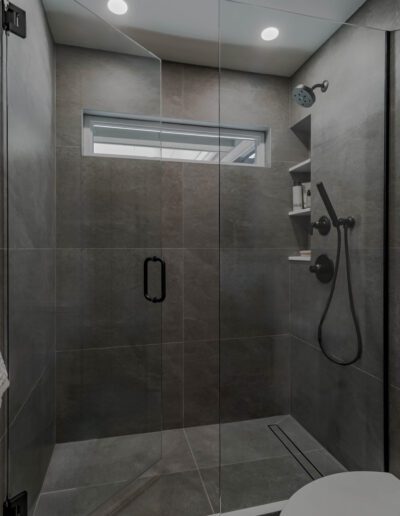 Modern bathroom featuring a glass shower enclosure, gray tiled walls, a handheld showerhead, and a small window for natural light.