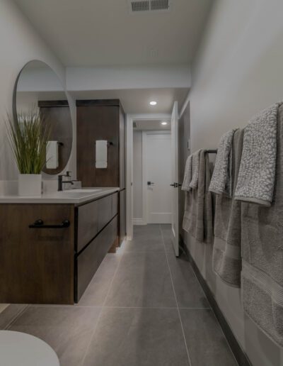 Modern bathroom interior featuring a dark wood vanity, round mirror, and gray towels hanging on the wall, with a view towards a white door at the end.