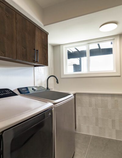 Modern laundry room with dark wood cabinets, a window, and stainless steel appliances including a washer and dryer.