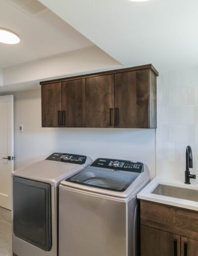 A modern laundry room featuring a stainless steel washer and dryer set, wooden cabinets, a sink, and a window.