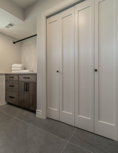 Modern bathroom featuring a gray tiled floor, a wooden vanity with dark cabinets, white towels stacked on a shelf, and three white doors with black handles.