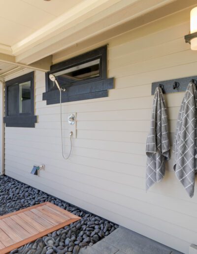 Exterior wall of a home featuring an outdoor shower with hanging gray towels, a wooden foot platform, and decorative rocks.