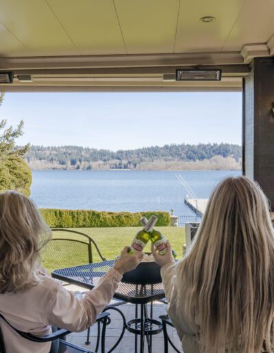 Two women toasting drinks on a patio overlooking a scenic lake.
